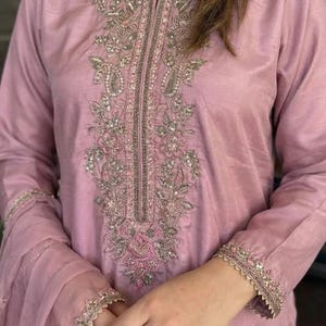 May include: A light mauve-colored tunic with intricate embroidery. The embroidery features floral and paisley designs with silver and pink accents. The sleeves and neckline are also embellished with delicate embroidery, creating an elegant look.