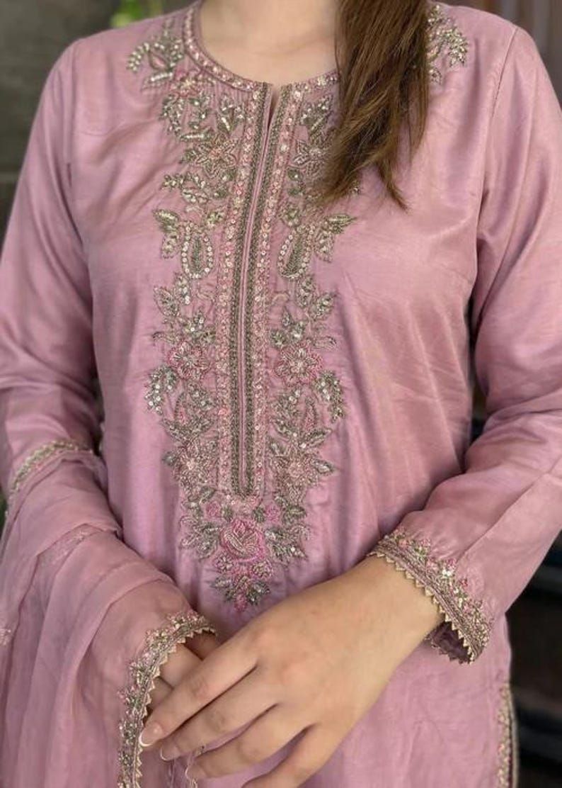 May include: A light mauve-colored tunic with intricate embroidery. The embroidery features floral and paisley designs with silver and pink accents. The sleeves and neckline are also embellished with delicate embroidery, creating an elegant look.