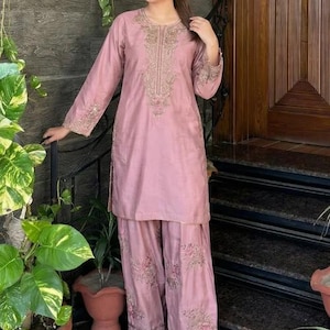 May include: A dusty rose-colored, embroidered kurta and palazzo pants set. The long-sleeved kurta features intricate gold embroidery around the neckline and cuffs. The palazzo pants have matching floral embroidery at the hem. The outfit is worn with embellished shoes.