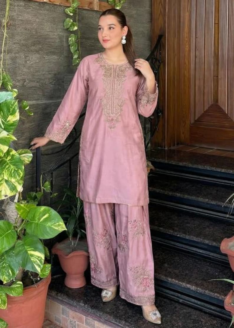 May include: A dusty rose-colored, embroidered kurta and palazzo pants set. The long-sleeved kurta features intricate gold embroidery around the neckline and cuffs. The palazzo pants have matching floral embroidery at the hem. The outfit is worn with embellished shoes.