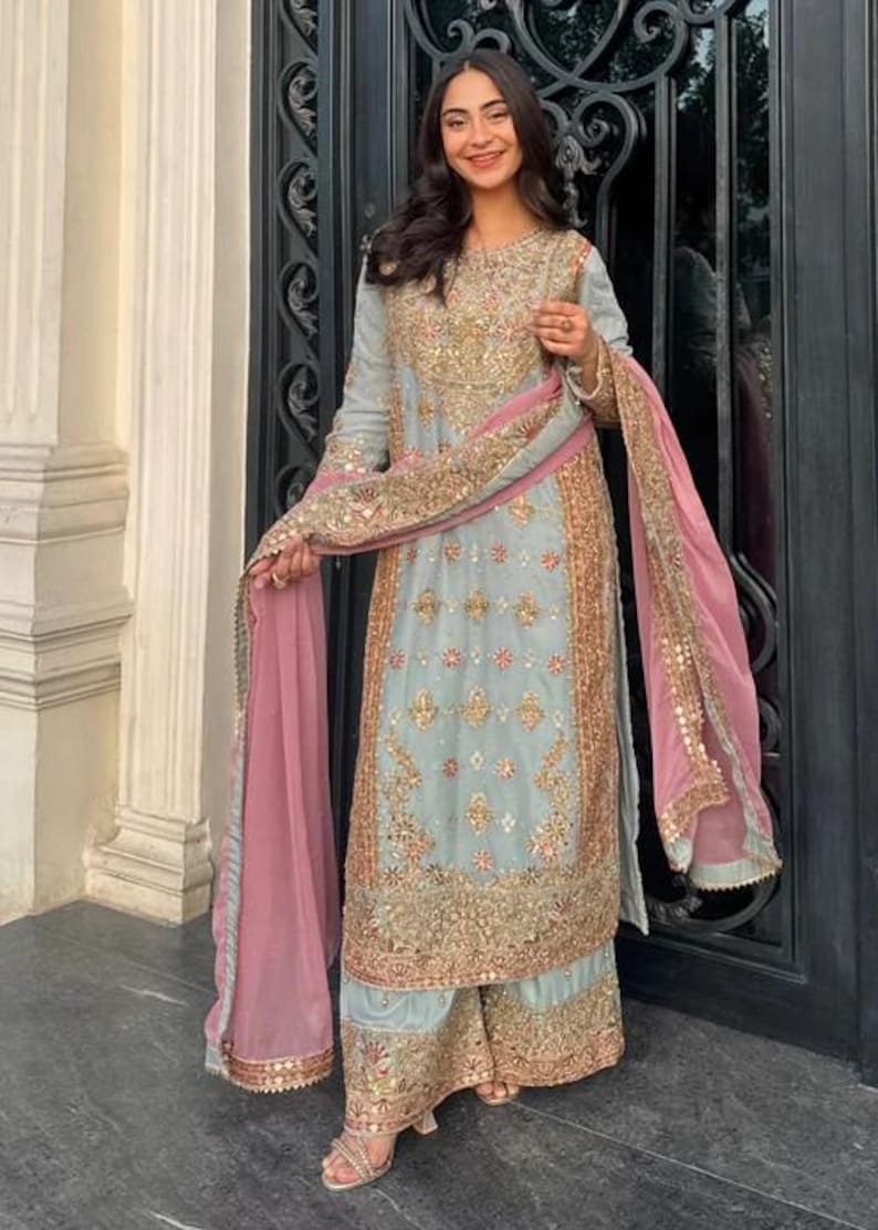 K&ouml;nnte beinhalten: Ein hellblaues Set aus lang&auml;rmeliger Tunika und weit geschnittener Hose mit aufwendigen Goldstickereien, Pailletten und Perlenarbeiten. Das Ensemble enth&auml;lt einen passenden hellblauen Dupatta mit rosa Rand und Goldbesatz &uuml;ber den Schultern.