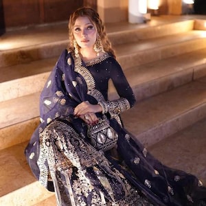 May include: A woman wearing a navy blue embroidered formal dress with gold accents, seated on stone steps. The dress features intricate patterns and a matching sheer shawl. She is also wearing large gold earrings and holding a small, embellished purse.