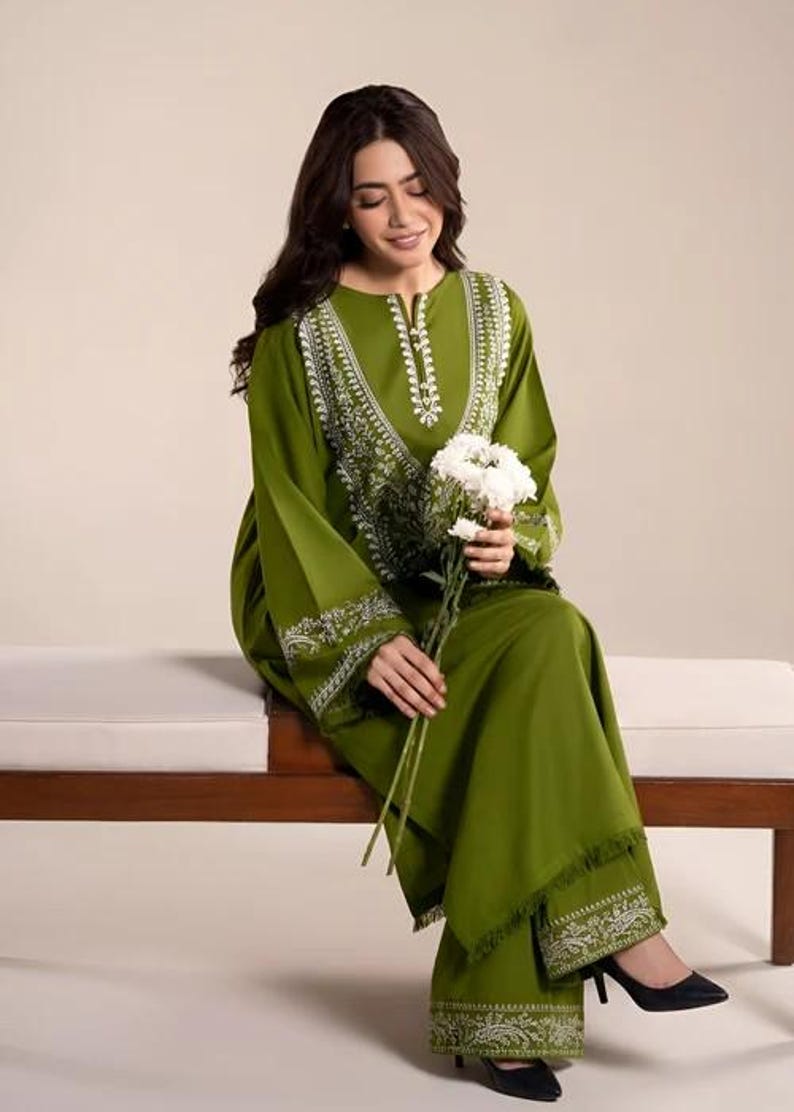 May include: Olive green two-piece outfit with white embroidered detailing. The long-sleeved top and wide-leg pants feature intricate patterns around the neckline, cuffs, and hem. The outfit is paired with black heels and a bouquet of white flowers.