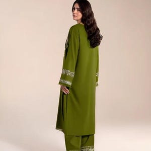 May include: Olive green two-piece outfit with white embroidered detailing. The long-sleeved tunic and matching pants feature intricate floral patterns along the cuffs and hem. The outfit is a solid color, with a simple, elegant design.