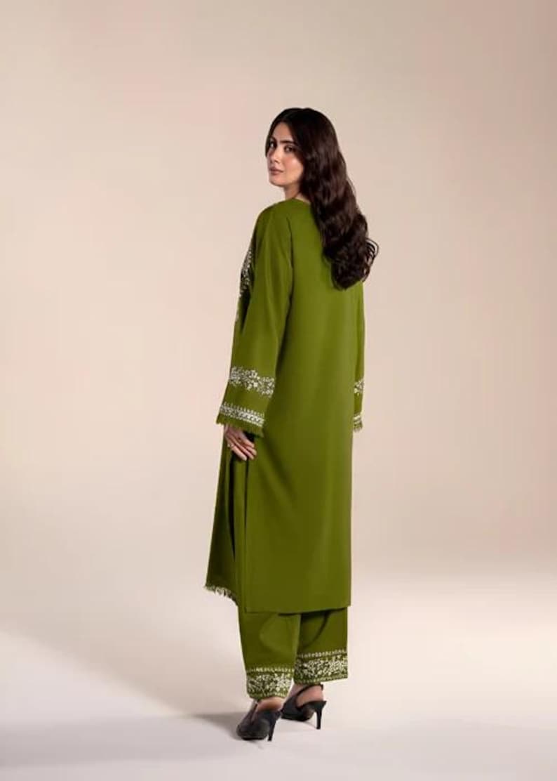 May include: Olive green two-piece outfit with white embroidered detailing. The long-sleeved tunic and matching pants feature intricate floral patterns along the cuffs and hem. The outfit is a solid color, with a simple, elegant design.