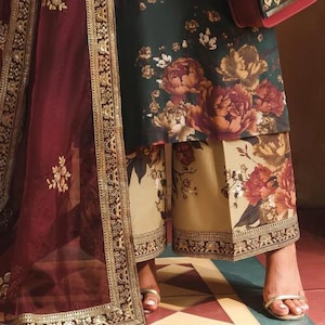May include: An ensemble featuring a dark green floral print top and beige pants with a matching floral design. A maroon sheer scarf with gold trim and a decorative purse with gold accents complete the look.