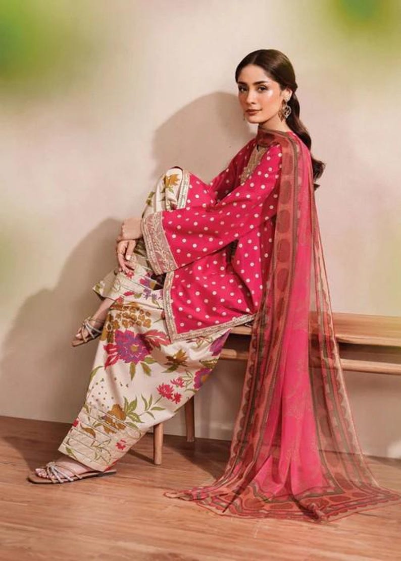 May include: A woman's ensemble featuring a pink polka-dot top with wide sleeves, paired with floral print pants. A matching sheer scarf drapes over the shoulder. The outfit is accented with gold trim.