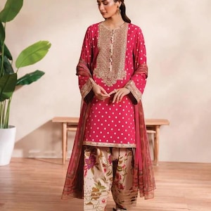 May include: A magenta-colored, embroidered tunic and matching dupatta set. The tunic features a polka dot pattern and gold embroidery around the neckline. The pants are a floral print. The outfit is paired with sandals.