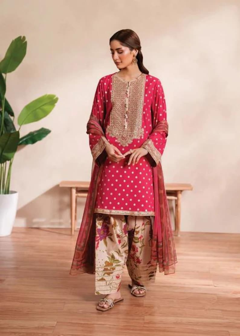 May include: A magenta-colored, embroidered tunic and matching dupatta set. The tunic features a polka dot pattern and gold embroidery around the neckline. The pants are a floral print. The outfit is paired with sandals.