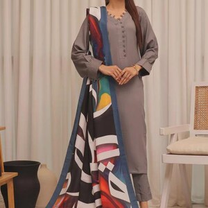 Puede incluir: Un conjunto salwar kameez gris con una parte superior de manga larga y pantalones a juego. El conjunto incluye una bufanda colorida con dise&ntilde;os abstractos en negro, blanco, azul y rojo. La parte superior tiene cuello en V y botones.