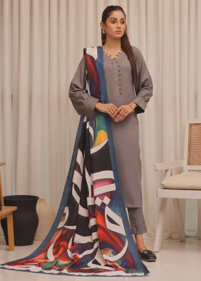 Puede incluir: Un conjunto salwar kameez gris con una parte superior de manga larga y pantalones a juego. El conjunto incluye una bufanda colorida con dise&ntilde;os abstractos en negro, blanco, azul y rojo. La parte superior tiene cuello en V y botones.