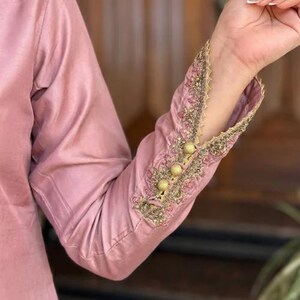 May include: A close-up of a dusty rose-colored long-sleeved garment. The sleeve cuff is intricately embroidered with gold and pink thread, featuring a scalloped edge and three gold buttons. The fabric has a subtle sheen.
