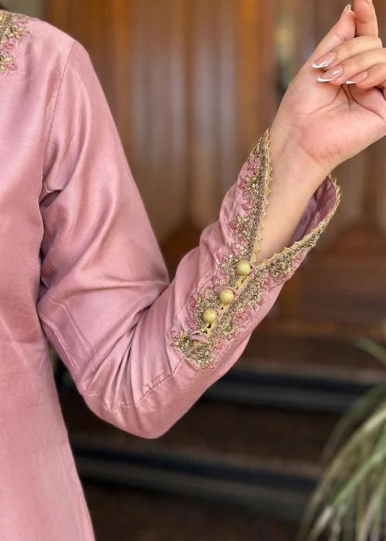 May include: A close-up of a dusty rose-colored long-sleeved garment. The sleeve cuff is intricately embroidered with gold and pink thread, featuring a scalloped edge and three gold buttons. The fabric has a subtle sheen.