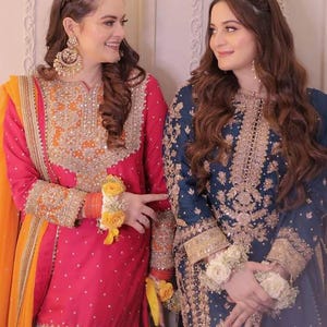 May include: Two women in ornate, embroidered traditional dresses. One wears a vibrant pink outfit with orange accents and gold embellishments. The other is in a deep blue dress with gold embroidery. Both outfits feature intricate detailing and floral wrist adornments.