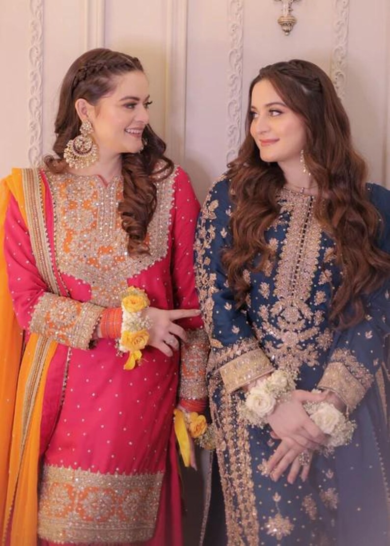 May include: Two women in ornate, embroidered traditional dresses. One wears a vibrant pink outfit with orange accents and gold embellishments. The other is in a deep blue dress with gold embroidery. Both outfits feature intricate detailing and floral wrist adornments.