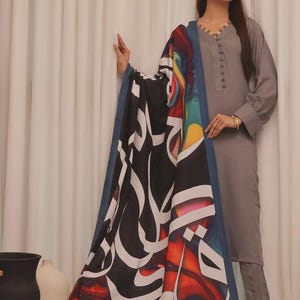 Puede incluir: Un conjunto gris de manga larga con una bufanda a juego. La bufanda presenta un fondo negro con caligraf&iacute;a blanca y dise&ntilde;os abstractos coloridos. El conjunto incluye una t&uacute;nica y pantalones.