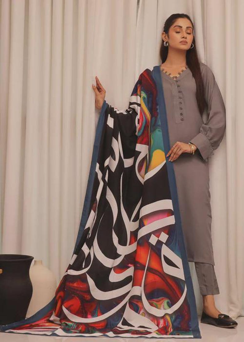 Puede incluir: Un conjunto gris de manga larga con una bufanda a juego. La bufanda presenta un fondo negro con caligraf&iacute;a blanca y dise&ntilde;os abstractos coloridos. El conjunto incluye una t&uacute;nica y pantalones.