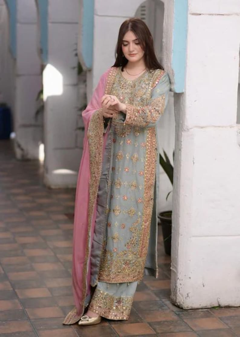 K&ouml;nnte beinhalten: Ein hellgraues Set aus einer lang&auml;rmeligen Tunika und einer weiten Hose mit aufwendigen Goldstickereien, Blumenmustern und kleinen Spiegelakzenten. Das Outfit wird durch ein langes, einfarbiges, rosafarbenes Dupatta erg&auml;nzt.