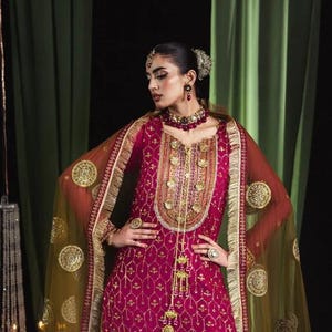 May include: A magenta-colored, embroidered formal dress with gold embellishments. The dress features a long-sleeved top and a matching bottom. A sheer, olive-green shawl with gold circular patterns drapes over the shoulders. The model is wearing matching jewelry.