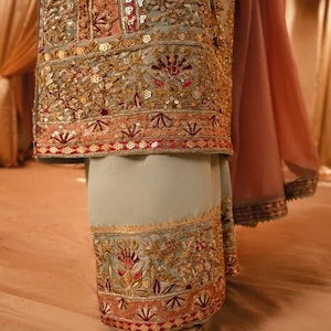 K&ouml;nnte beinhalten: Ein salbeigr&uuml;nes Tunika- und Hosen-Set mit weiter Passform, verziert mit aufwendigen Goldstickereien, Pailletten und floralen Motiven in Korallen- und Dunkelrott&ouml;nen. Das Kleidungsst&uuml;ck umfasst einen flie&szlig;enden, transparenten korallenfarbenen Stoff.