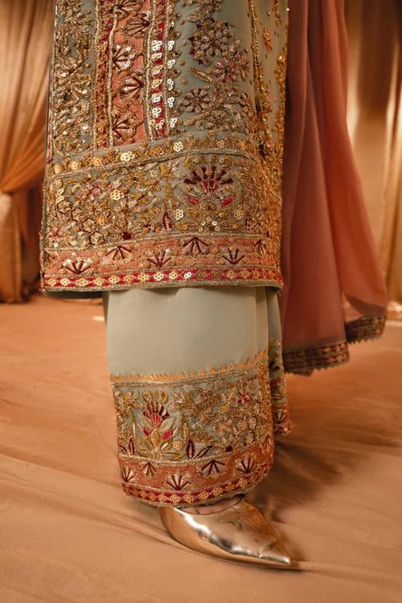K&ouml;nnte beinhalten: Ein salbeigr&uuml;nes Tunika- und Hosen-Set mit weiter Passform, verziert mit aufwendigen Goldstickereien, Pailletten und floralen Motiven in Korallen- und Dunkelrott&ouml;nen. Das Kleidungsst&uuml;ck umfasst einen flie&szlig;enden, transparenten korallenfarbenen Stoff.