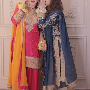 May include: Two women in traditional South Asian attire. One wears a vibrant pink outfit with gold embroidery and an orange scarf. The other is in a navy blue ensemble with gold accents. Both outfits feature intricate detailing.