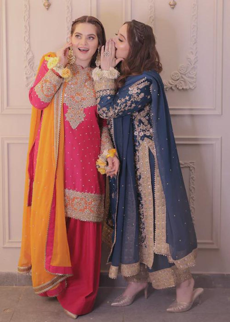 May include: Two women in traditional South Asian attire. One wears a vibrant pink outfit with gold embroidery and an orange scarf. The other is in a navy blue ensemble with gold accents. Both outfits feature intricate detailing.