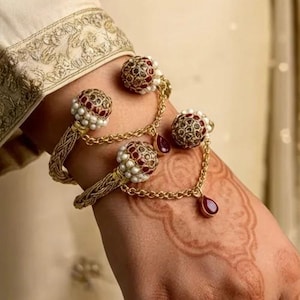 May include: A gold-toned bracelet with intricate detailing. The bracelet features decorative spheres with red and white accents, connected by delicate chains. A teardrop-shaped red gem hangs from the chain. The bracelet is worn on a wrist with henna designs.