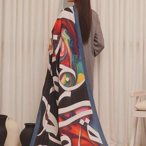 Puede incluir: Una bufanda larga y rectangular con un vibrante dise&ntilde;o abstracto en rojo, azul, amarillo y verde. La bufanda presenta una gran caligraf&iacute;a &aacute;rabe blanca sobre un fondo negro, enmarcada por un borde azul. La bufanda est&aacute; drapeada sobre una persona.