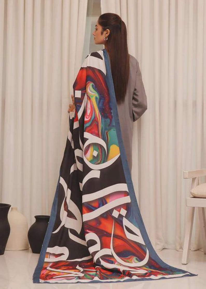 Puede incluir: Una bufanda larga y rectangular con un vibrante dise&ntilde;o abstracto en rojo, azul, amarillo y verde. La bufanda presenta una gran caligraf&iacute;a &aacute;rabe blanca sobre un fondo negro, enmarcada por un borde azul. La bufanda est&aacute; drapeada sobre una persona.