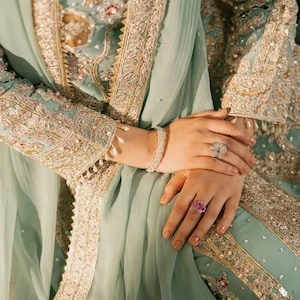 May include: A light mint green long-sleeved garment featuring intricate gold embroidery, silver beadwork, and sequins. The sleeves are adorned with dangling pearl-like accents. A silver bracelet and two rings, one floral-shaped and one pink, are worn.