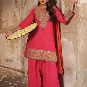 May include: A vibrant pink salwar kameez ensemble with intricate gold embroidery. The outfit includes a long-sleeved tunic, wide-legged pants, and an orange scarf. The tunic is adorned with small, sparkling embellishments.