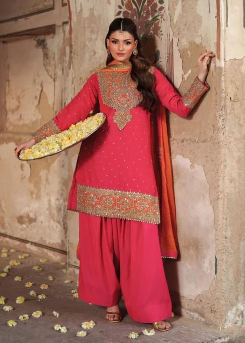 May include: A vibrant pink salwar kameez ensemble with intricate gold embroidery. The outfit includes a long-sleeved tunic, wide-legged pants, and an orange scarf. The tunic is adorned with small, sparkling embellishments.