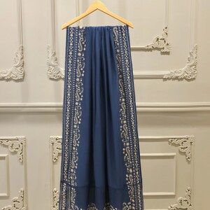 May include: A navy blue scarf with intricate white floral embroidery along the edges. The scarf is hanging on a wooden hanger against a neutral wall. The embroidery features delicate floral and leaf patterns.