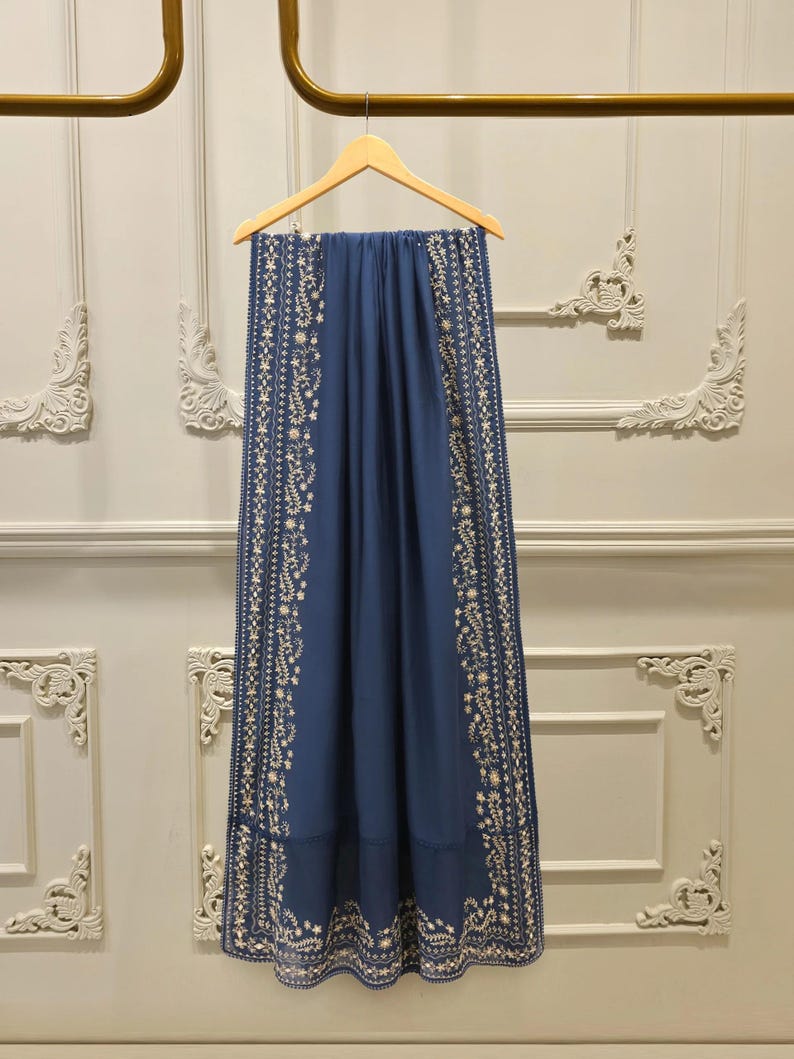 May include: A navy blue scarf with intricate white floral embroidery along the edges. The scarf is hanging on a wooden hanger against a neutral wall. The embroidery features delicate floral and leaf patterns.