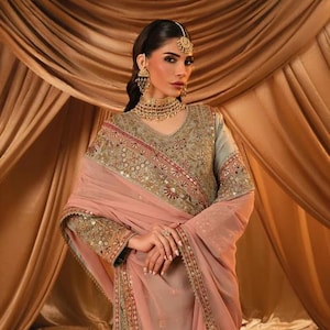 K&ouml;nnte beinhalten: Eine salbeigr&uuml;ne Tunika mit aufwendiger Goldstickerei und Spiegelarbeit, kombiniert mit einer zart pfirsichfarbenen Chiffon-Dupatta. Das Set umfasst eine passende goldfarbene Halskette, h&auml;ngende Ohrringe und einen Kopfschmuck mit klaren Steinen.