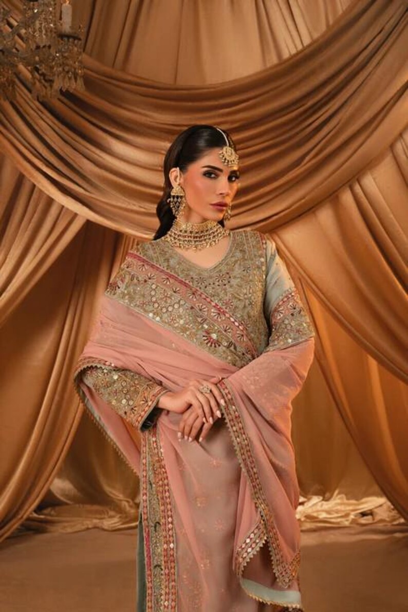 K&ouml;nnte beinhalten: Eine salbeigr&uuml;ne Tunika mit aufwendiger Goldstickerei und Spiegelarbeit, kombiniert mit einer zart pfirsichfarbenen Chiffon-Dupatta. Das Set umfasst eine passende goldfarbene Halskette, h&auml;ngende Ohrringe und einen Kopfschmuck mit klaren Steinen.