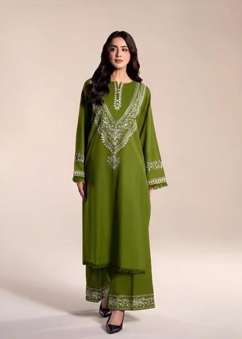 May include: Olive green embroidered tunic and matching pants. The long-sleeved tunic features intricate white embroidery around the neckline, cuffs, and hem. The pants have a matching embroidered design at the hem. The outfit is a coordinated set.