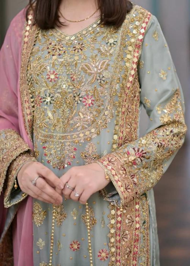 K&ouml;nnte beinhalten: Eine salbeigr&uuml;ne Tunika mit langen &Auml;rmeln, verziert mit aufwendigen Goldstickereien, Pailletten und floralen Motiven in Rot und Pink. Das Kleidungsst&uuml;ck hat einen V-Ausschnitt und wird mit einem transparenten, rosafarbenen Dupatta mit Goldborte kombiniert.