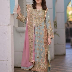 K&ouml;nnte beinhalten: Ein hellblaues Set aus lang&auml;rmeliger Tunika und weit geschnittener Hose mit aufwendigen Goldstickereien, Pailletten und Spiegelarbeiten. Ein passender hellblauer Dupatta mit breitem, goldbesticktem Rand und einem rosafarbenen Einsatz liegt &uuml;ber der Schulter.