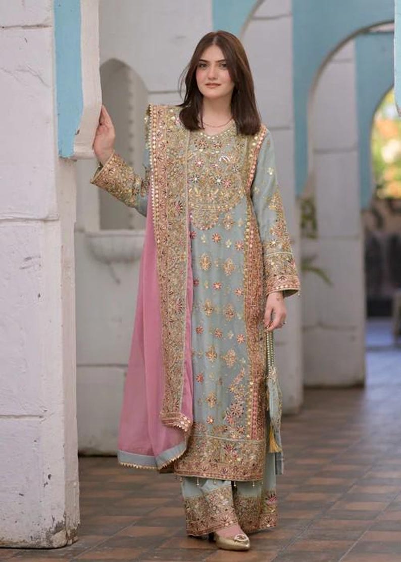 K&ouml;nnte beinhalten: Ein hellblaues Set aus lang&auml;rmeliger Tunika und weit geschnittener Hose mit aufwendigen Goldstickereien, Pailletten und Spiegelarbeiten. Ein passender hellblauer Dupatta mit breitem, goldbesticktem Rand und einem rosafarbenen Einsatz liegt &uuml;ber der Schulter.