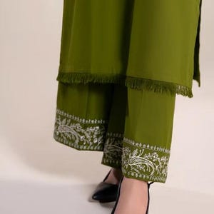 May include: Olive green two-piece outfit featuring a long tunic with fringe trim and matching pants. The pants have intricate white floral embroidery along the hem. Black pointed-toe heels complete the ensemble.