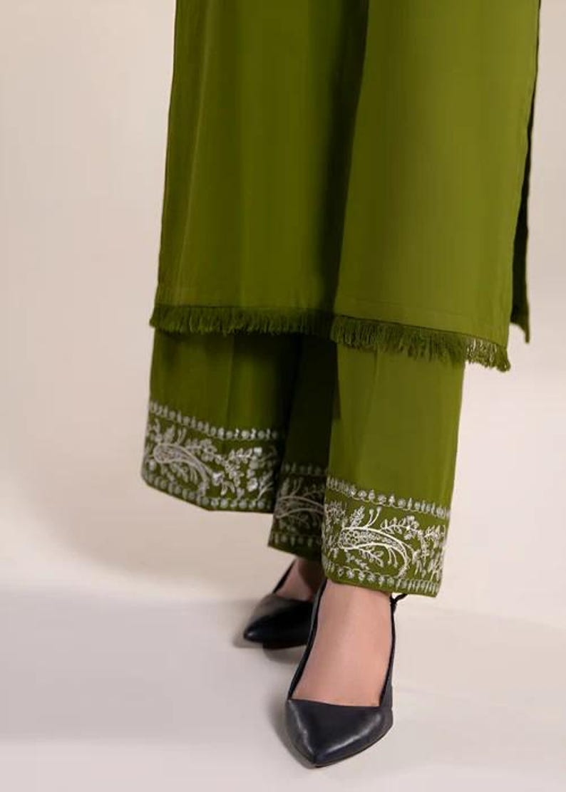 May include: Olive green two-piece outfit featuring a long tunic with fringe trim and matching pants. The pants have intricate white floral embroidery along the hem. Black pointed-toe heels complete the ensemble.