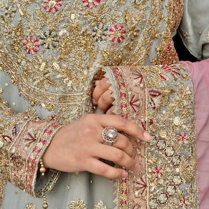 May include: Close-up of a light blue embroidered garment with intricate gold and red floral patterns. The sleeve cuff and neckline feature detailed embroidery. A large, ornate ring with a central stone is visible on the hand.