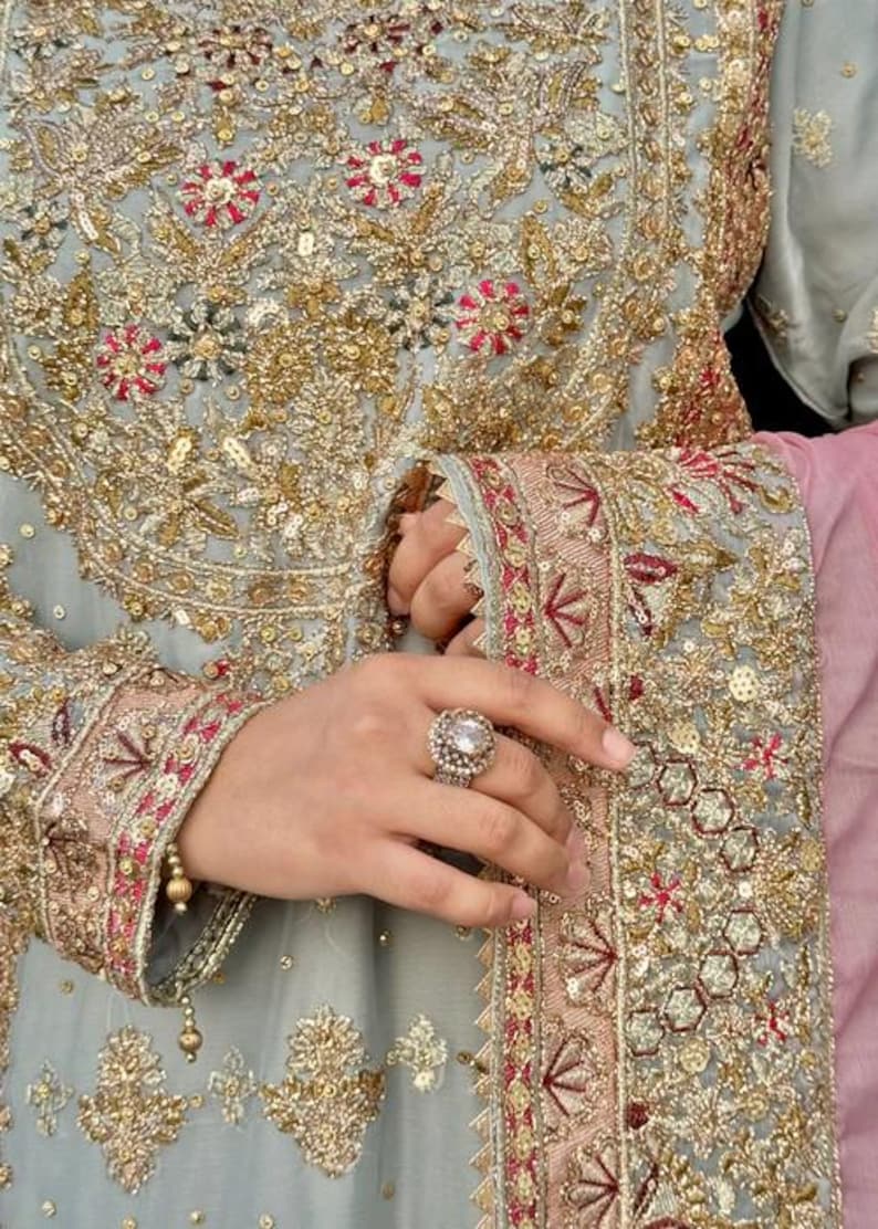 K&ouml;nnte beinhalten: Ein hellgraues Kleidungsst&uuml;ck mit aufwendigen Goldstickereien, Pailletten und kleinen rosa Blumenakzenten. Der Stoff ist reich mit Metallf&auml;den verziert. Ein Ring mit einem gro&szlig;en, runden, klaren Stein und einer dekorativen Fassung wird am Finger getragen.