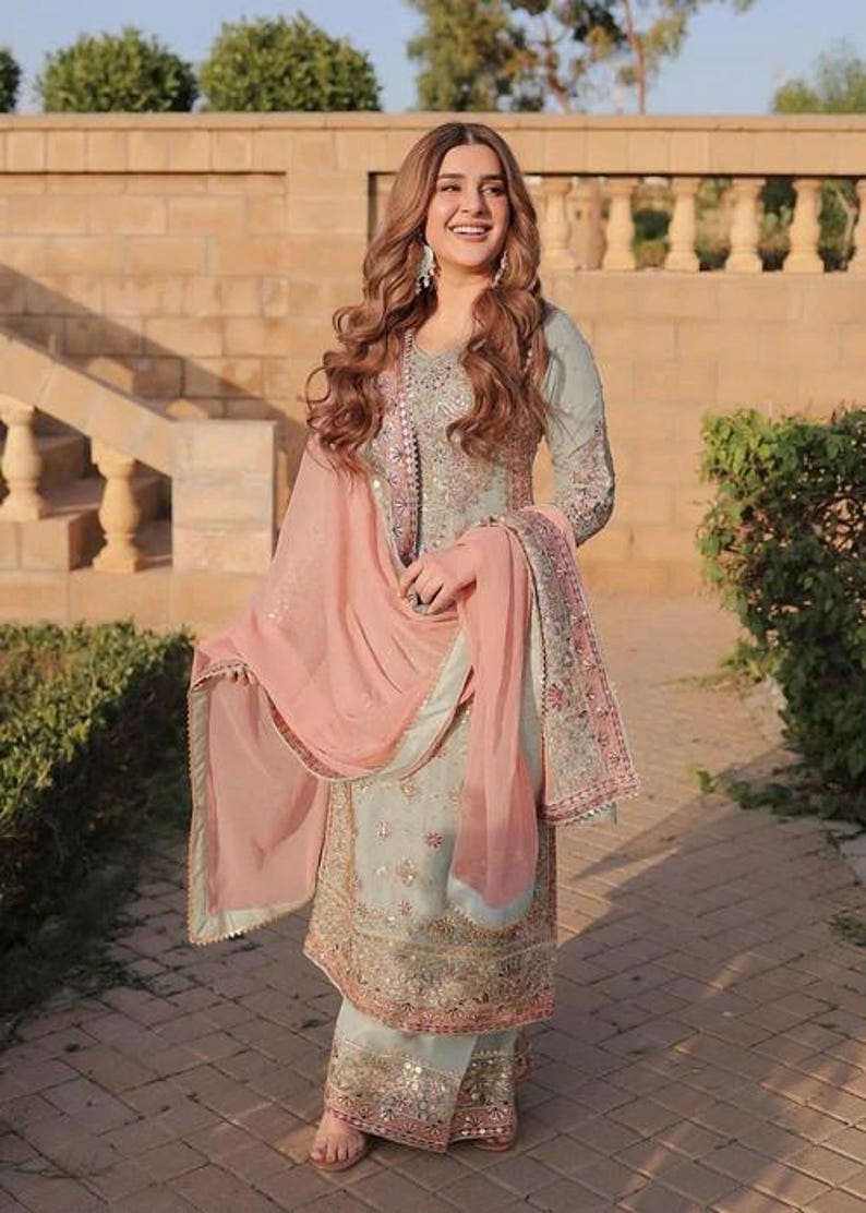 May include: A light blue and pink embroidered salwar kameez. The outfit features intricate gold and silver embellishments. A matching pink dupatta is draped over the shoulders. The ensemble is worn outdoors.