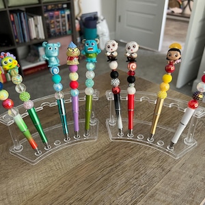May include: A collection of decorative pens with character-themed toppers. Each pen features a unique design with colorful beads and a character figurine. The pens are displayed in a clear acrylic holder on a wooden surface.