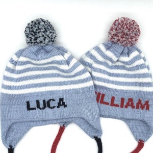 May include: Two blue and white striped knit hats with ear flaps and pom-poms. One hat has the name "LUCA" in black letters, and the other has "WILLIAM" in red letters. The hats have black and red ties.