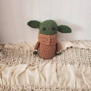 May include: A green and brown crocheted plush toy resembling a character from the Star Wars franchise. The toy has large eyes and a brown crocheted collar.