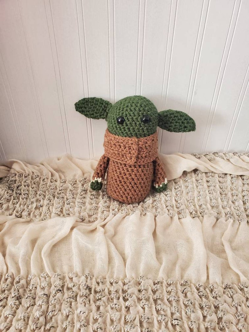 May include: A green and brown crocheted plush toy resembling a character from the Star Wars franchise. The toy has large eyes and a brown crocheted collar.